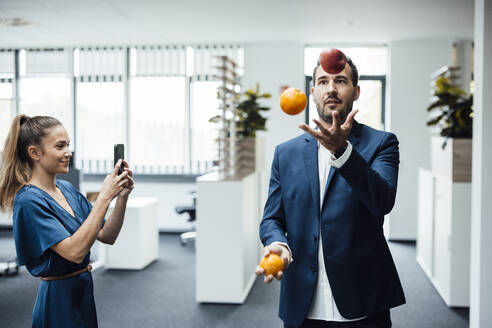 Junge Geschäftsfrau fotografiert Kollegin beim Jonglieren von Obst mit dem Smartphone im Büro - JOSEF17514