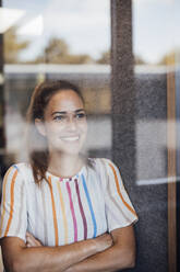 Lächelnde Geschäftsfrau mit verschränkten Armen durch Glas gesehen - JOSEF17546