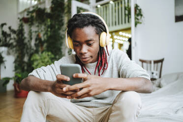 Junger Mann mit Kopfhörern, der zu Hause sein Smartphone benutzt - MDOF00756