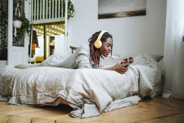 Smiling young man wearing headphones and using smart phone lying on bed at home - MDOF00759