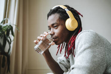 Nachdenklicher junger Mann mit Kopfhörern trinkt zu Hause Wasser - MDOF00768
