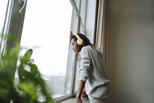 Junger Mann mit drahtlosen Kopfhörern, der Musik hört und durch das Fenster seines Hauses schaut - MDOF00770