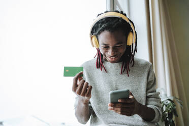 Lächelnder Mann mit Kreditkarte und Smartphone zu Hause - MDOF00771