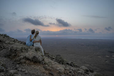 Paar betrachtet den Sonnenuntergang auf einem Berg sitzend - FBAF02118