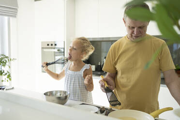 Girl licking egg beater by grandfather preparing food in kitchen - SVKF01323