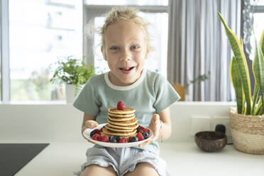Happy girl holding plate of fresh pancakes with berries in kitchen at home - SVKF01329