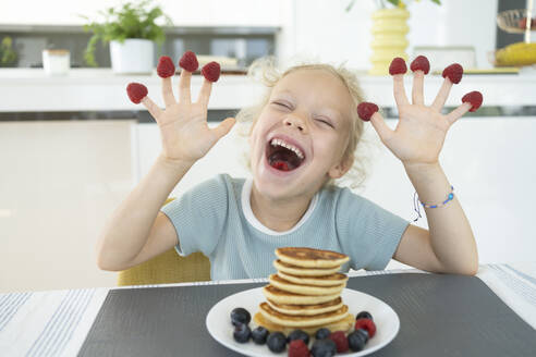 Playful girl with raspberries on fingers laughing at dining table at home - SVKF01331