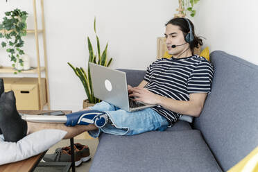 Freelancer with prosthetic leg using laptop wearing headset sitting on sofa at home - JCZF01208