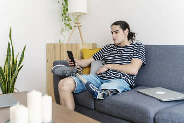 Young man with prosthetic leg using smart phone on sofa at home - JCZF01211