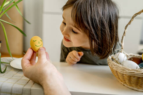 Lächelnder Junge betrachtet ein dekoriertes Osterei zu Hause - ANAF01049