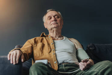 Älterer Mann mit geschlossenen Augen, der auf dem Sofa sitzend seinen Blutdruck kontrolliert - VSNF00563
