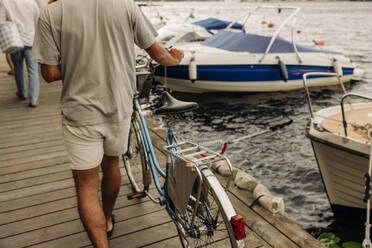 Rückansicht eines Mannes, der ein Fahrrad auf einem Pier im Hafen schiebt - MASF35442