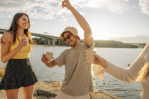 Unbekümmerter junger Mann tanzt mit erhobener Hand von Freundinnen beim Picknick - MASF35449