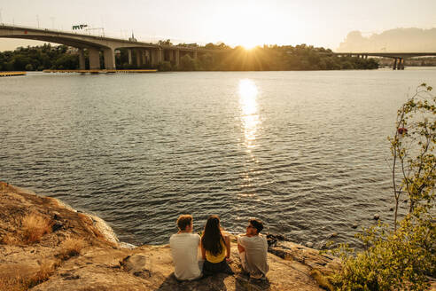 Hohe Winkel Ansicht von Freunden sitzen und reden auf Felsen in der Nähe von Meer bei Sonnenuntergang - MASF35454