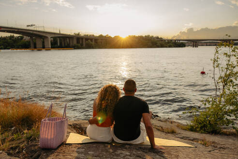 Rückansicht eines Paares mit Blick auf den Sonnenuntergang, das auf einer Picknickdecke am Meer sitzt - MASF35456