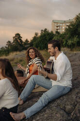 Lächelnder junger Mann spielt Gitarre, während er mit Freunden beim Picknick auf einem Felsen sitzt - MASF35459