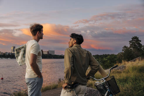 Seitenansicht eines jungen Mannes im Gespräch mit einem männlichen Freund mit Fahrrad bei Sonnenuntergang - MASF35464