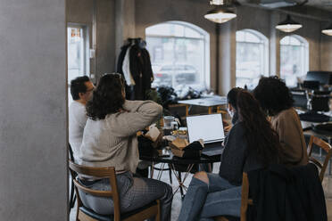 Junger Programmierer bei der Arbeit am Laptop mit männlichen und weiblichen Kollegen beim Mittagessen in einem Start-up-Unternehmen - MASF35480