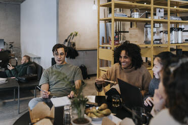 Lächelnde Geschäftskollegen, die beim Mittagessen am Tisch in einem kreativen Büro diskutieren - MASF35481