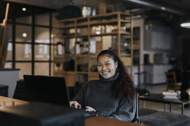 Portrait of happy businesswoman with laptop at desk in creative office - MASF35505