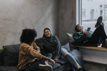 Fröhliche Geschäftsfrauen diskutieren auf dem Sofa mit einem männlichen Kollegen, der ein Mobiltelefon im Büro benutzt - MASF35522