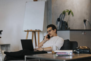 Junger männlicher Unternehmer, der mit seinem Smartphone telefoniert, während er an einem Laptop in einem kreativen Büro arbeitet - MASF35557