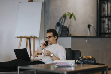 Geschäftsmann, der einen Kaffee trinkt, während er mit seinem Smartphone telefoniert und an seinem Laptop im Büro arbeitet - MASF35558