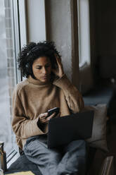 Female entrepreneur wearing wireless headphones and working on laptop by window in creative office - MASF35561