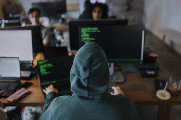 Rückansicht eines jungen männlichen Hackers mit Kapuze, der an einem Schreibtisch in einer kreativen Arbeitsumgebung am Computer programmiert - MASF35594