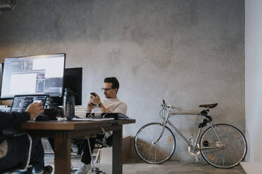 Geschäftsmann, der ein Smartphone benutzt, während ein männlicher Kollege an einem Laptop und Computer am Arbeitsplatz arbeitet - MASF35605