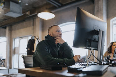 Junger männlicher Hacker mit Hand am Kinn, der einen Computer am Schreibtisch in einem kreativen Büro benutzt - MASF35606