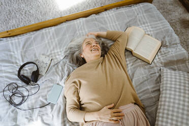 Happy senior woman with book and headphones lying down on bed - MASF35635