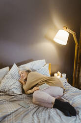 Ältere Frau schläft auf dem Bett in der Nähe einer beleuchteten Lampe zu Hause - MASF35637