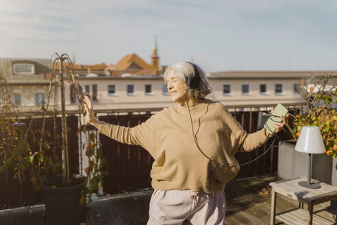 Sorglose ältere Frau, die auf der Terrasse stehend Musik über Kopfhörer hört - MASF35638