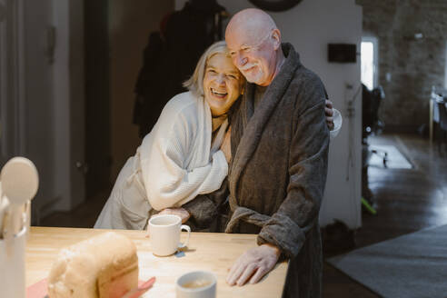 Happy senior woman embracing man wearing bathrobe at home - MASF35644