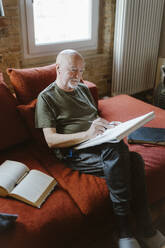 Senior man doing painting while sitting on sofa by book at home - MASF35654