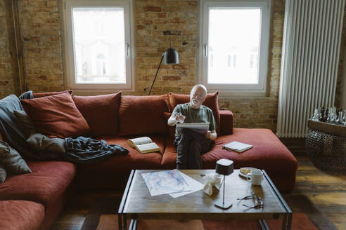 Älterer Mann malt, während er auf dem Sofa im Wohnzimmer zu Hause sitzt - MASF35655
