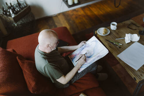 High angle view of senior man painting face over paper while sitting on sofa at home - MASF35656