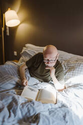 Älterer Mann mit Hand am Kinn liest ein Buch, während er zu Hause auf dem Bett liegt - MASF35659