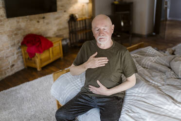 Älterer Mann mit geschlossenen Augen macht Atemübungen, während er zu Hause auf dem Bett sitzt - MASF35661