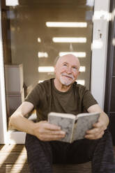 Lächelnder älterer Mann, der mit einem Buch zu Hause sitzt und träumt - MASF35663