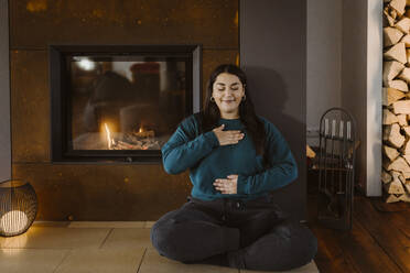 Woman with eyes closed doing breathing exercise while sitting cross-legged near fireplace at home - MASF35670
