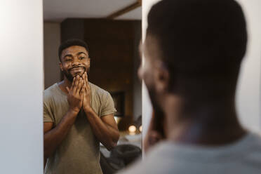 Smiling young man touching face while looking in mirror at home - MASF35689