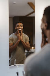 Smiling man applying moisturizer while looking in mirror at home - MASF35691