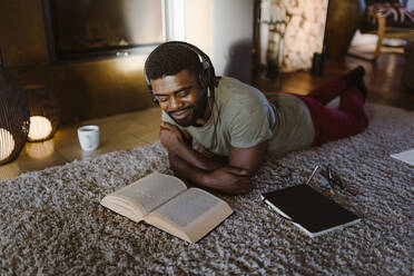 Smiling young man listening to music and reading book while lying on carpet at home - MASF35695