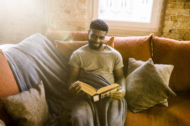 Lächelnder junger Mann liest ein Buch, während er zu Hause auf dem Sofa mit einer Decke sitzt - MASF35697