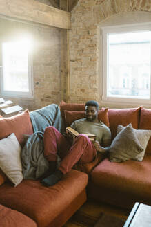 Junger Mann liest ein Buch und entspannt sich auf dem Sofa zu Hause - MASF35699