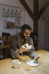 Smiling man using smart phone and having doughnut at home - MASF35701