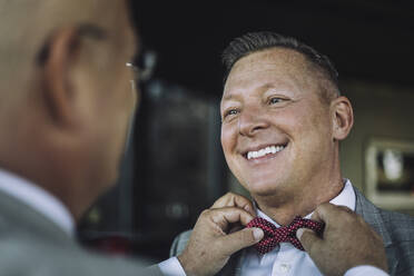 Smiling gay man looking at partner adjusting bow tie before wedding - MASF35703