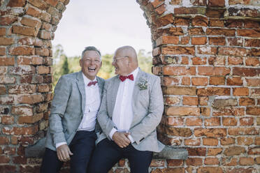 Gut gekleidetes glückliches schwules Paar, das während der Hochzeit am Fenster sitzt - MASF35708